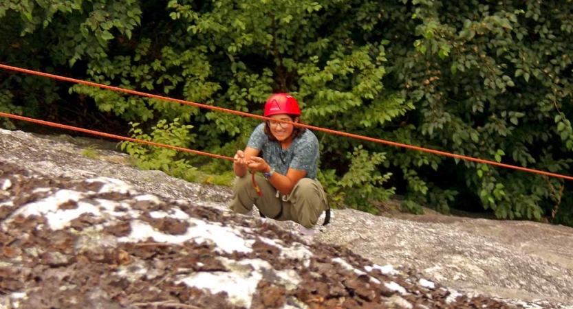 A person wearing a red helmet is attached to ropes by safety gear as they crouch on a rock wall while climbing up. 